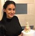 A woman with long dark hair smiles while holding a decorated cake with a small figure on top, indoors.