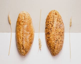 Two loaves of bread, one lighter and one darker with seeds, positioned vertically with a wheat stalk on each side against a two-tone backdrop, evoke the comforting essence of home.