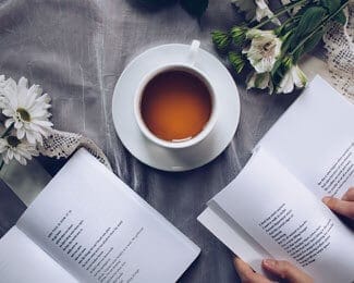 A cup of tea and two open books on a gray tablecloth, surrounded by white flowers, create a cozy home atmosphere. A pair of hands is holding one of the books.
