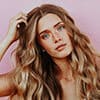 A woman with long, wavy hair and natural makeup poses against a pink background, gazing at the camera with her hand in her hair, exuding a sense of home and comfort.