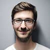 A man with short brown hair, beard, and eyeglasses, wearing a white t-shirt, smiles warmly against a gray background, giving off a comfortable home vibe.