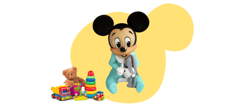 A fondant cake topper featuring Baby Mickey Mouse in pajamas holding a blanket, sitting beside colorful toys, including a teddy bear, stacked rings, and vehicles.