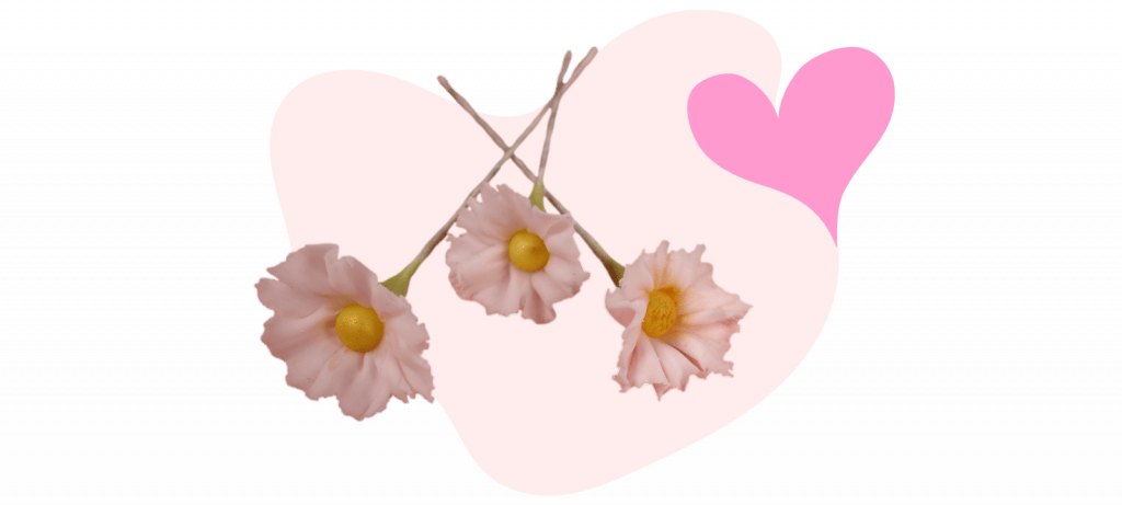 Three pink fondant flowers with yellow centers are arranged in front of a pink and white abstract background featuring a heart shape.
