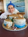 A person wearing a blue bandana smiles behind a beach-themed wedding cake with a "Married" sign and a charming cake topper among the beach decorations.