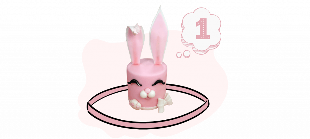 Pink cake decorated with bunny ears, eyes, and a nose, surrounded by candy. Number "1" inside a thought bubble. Ideal for showcasing your fondant cake decoration skills or following along with a video tutorial.