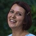 A woman with short brown hair and a hairpin is smiling warmly against a blurred green natural background, resembling a charming cake topper.