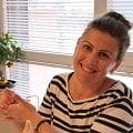 A smiling woman with dark hair in a bun wearing a striped shirt sits indoors with a plant and window blinds in the background, looking as charming as a cake topper.