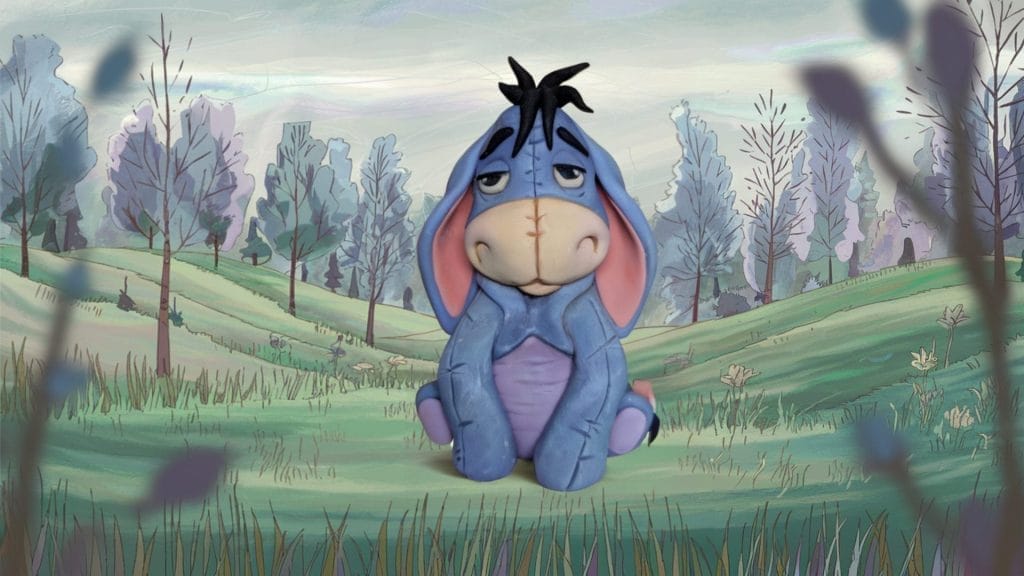 A cake topper Eeyore sits on green grass in a cartoon-style landscape with trees and hills under a cloudy sky.